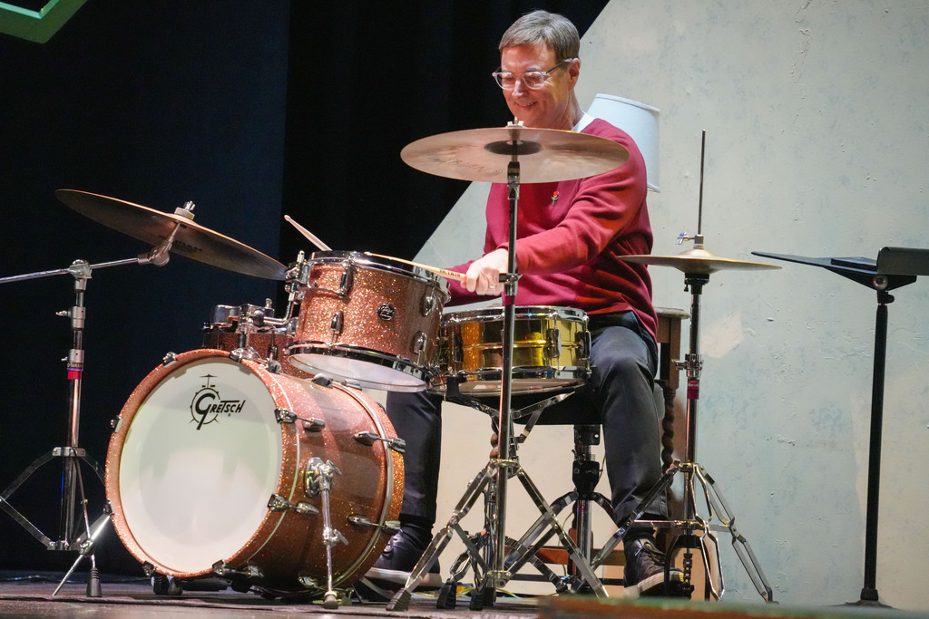 Person in a red long-sleeve shirt playing a drum set with cymbals on a stage.