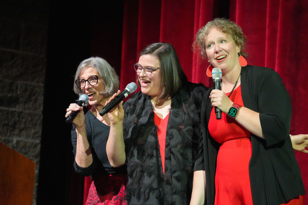Three women on stage with microphones, smiling. One wears a red dress, another black, and the third has a red curtain in the background.