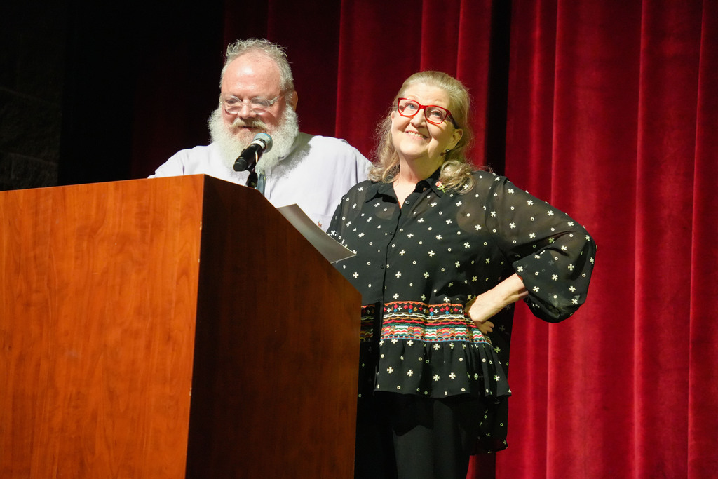 Two people at a podium with a microphone in front of a red curtain backdrop.