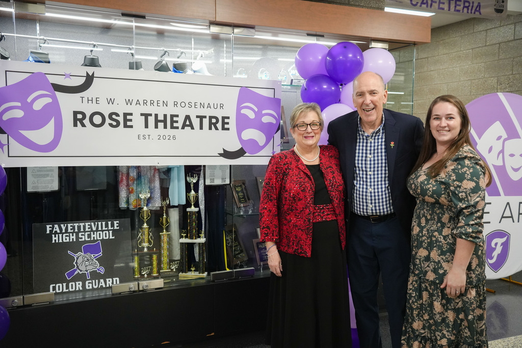 Three people stand before a theatre entrance. A sign reads "The W. Warren Rosenau Rose Theatre." Purple balloons are in the background.