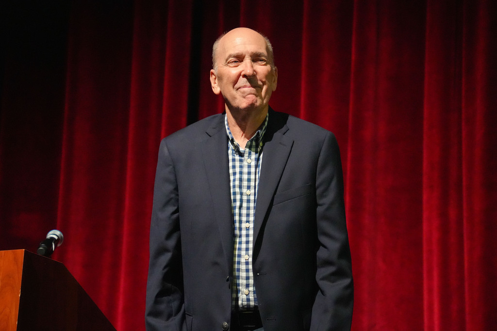 Man wearing a black blazer over a blue and white checkered shirt standing on stage with a microphone.