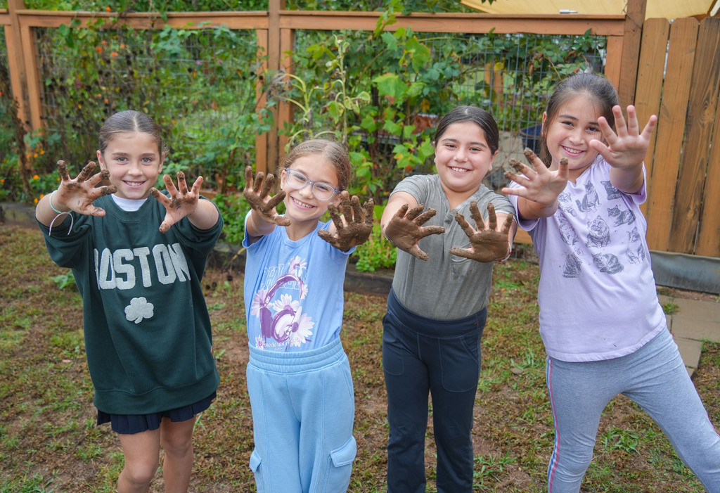 Four girls with dirty hands stand in a garden. Behind them, a wooden fence encloses plants and trees.