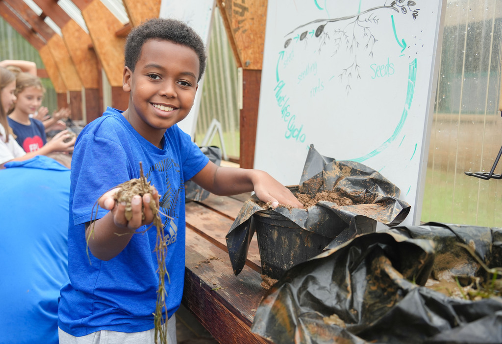 A smiling child holds a plant outdoors, near a wooden desk and green bags. Behind, others observe.