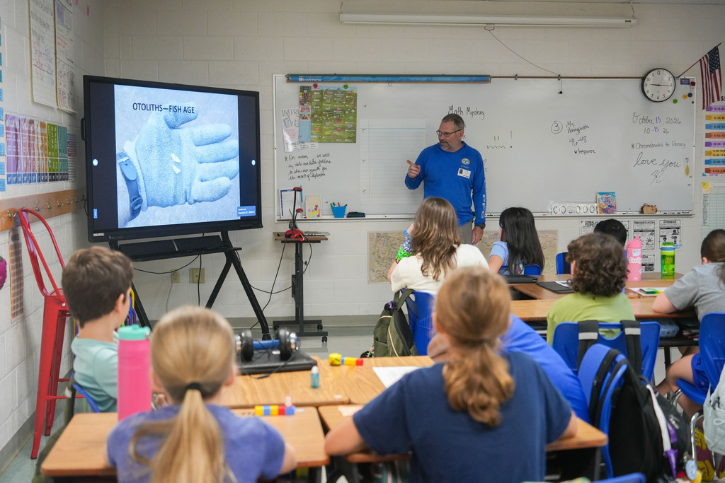 A teacher in blue stands before a screen showing a hand, instructing students seated at desks.