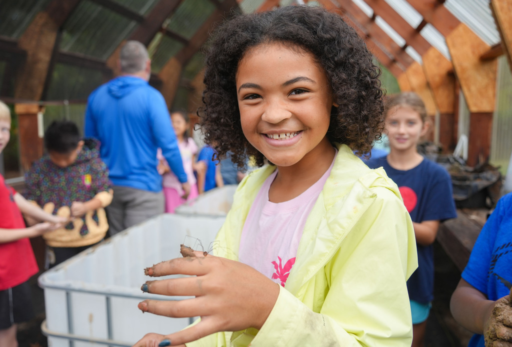 A girl with curly hair smiles and holds a bug, wearing a yellow jacket. Behind her, several children and adults are in a greenhouse.