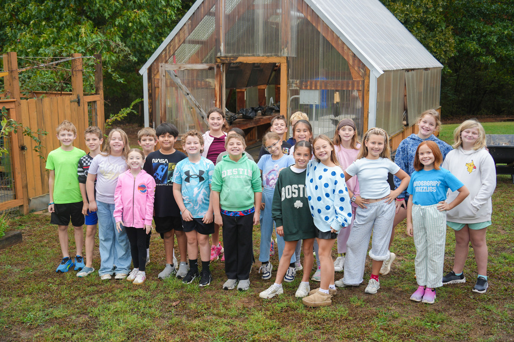 Group of children in various tops and pants posing in front of a small greenhouse.