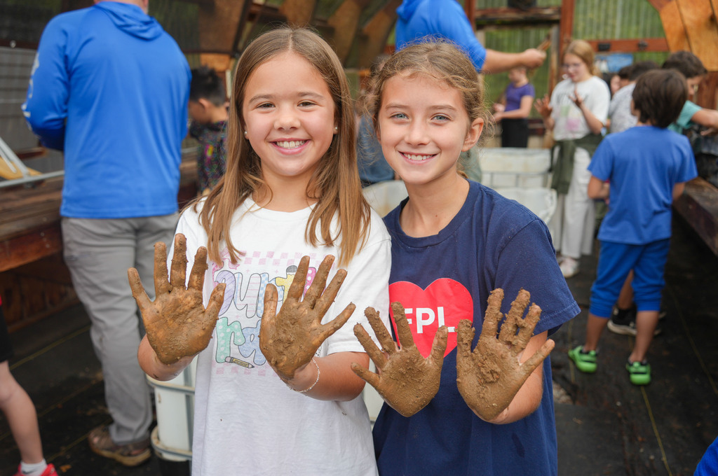 Two girls with mud on their hands smile at the camera in an outdoor area with others.