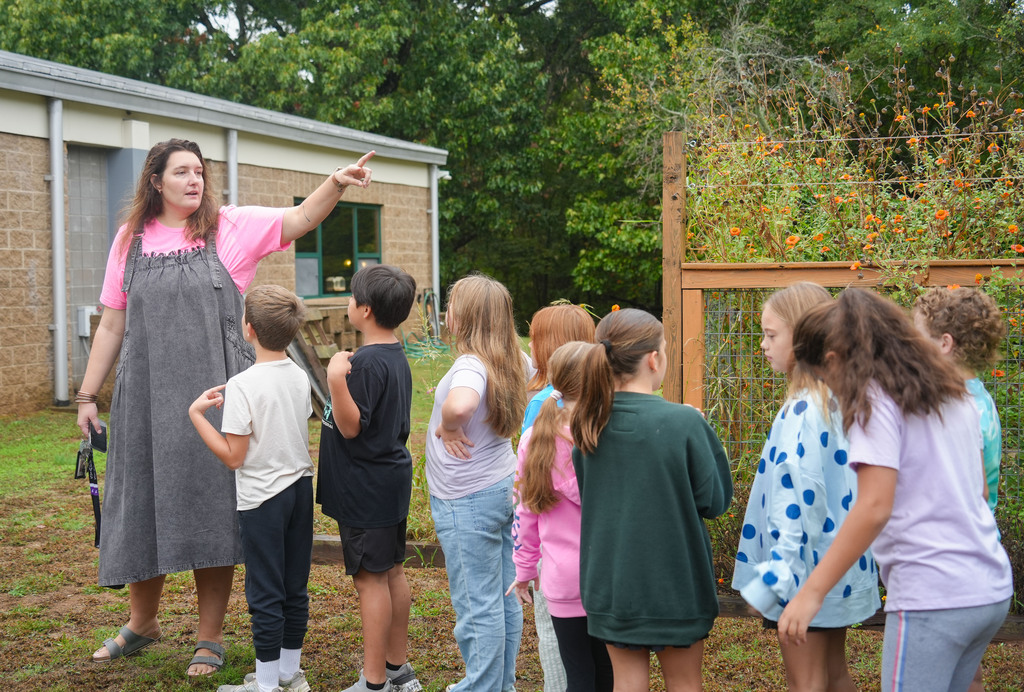 An adult woman points at a garden with plants. A group of children, all wearing different colored shirts, listen.