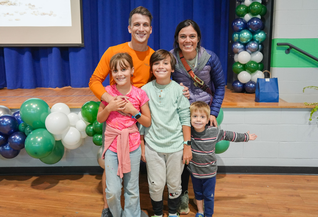 A family of five stands on a stage. Three children hold hands. Two adults stand behind the children. Green and white balloons are in the background.