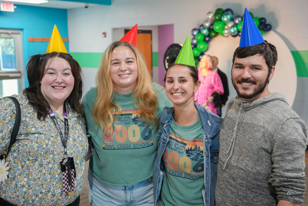 Four people in matching green shirts pose for a photo with party hats in a colorful room.