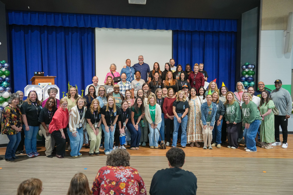 A group of people in various clothing and footwear standing on a stage in front of blue curtains.