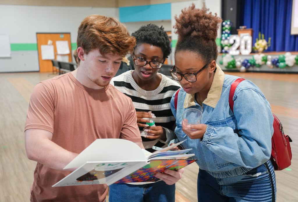 Three students examine a colorful book; one holds a water bottle, another a cup, and the third has a backpack.