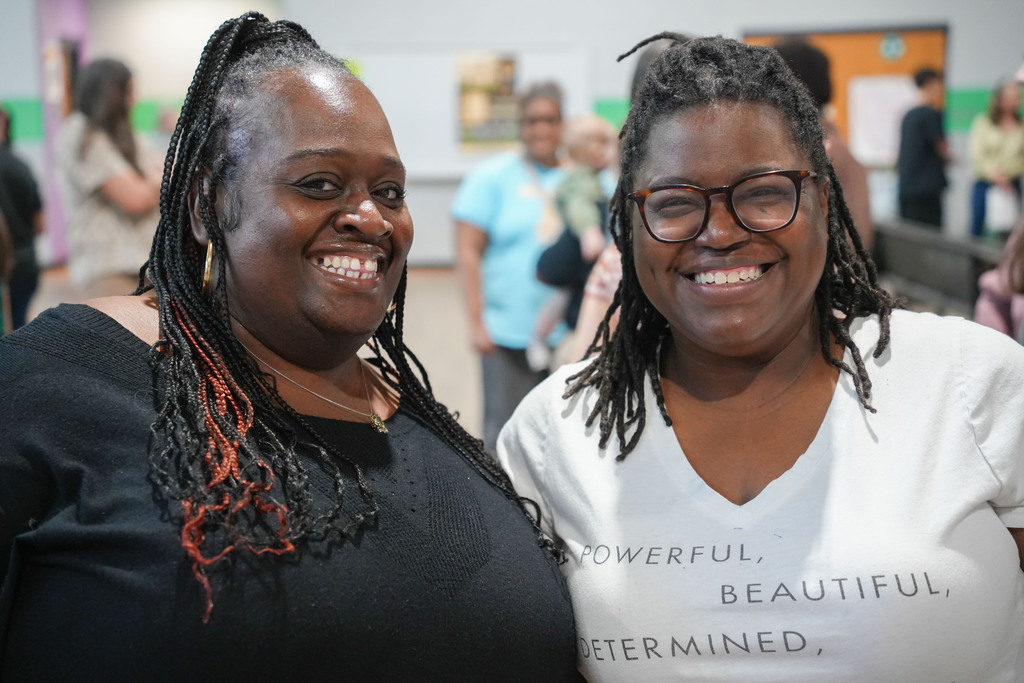 Two women smiling, standing next to each other. One wears a black top; the other wears a white t-shirt.