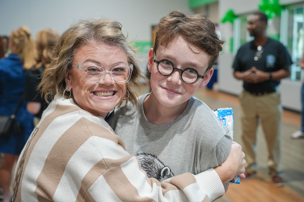 A smiling adult woman with glasses hugs a child with glasses in a room with others.