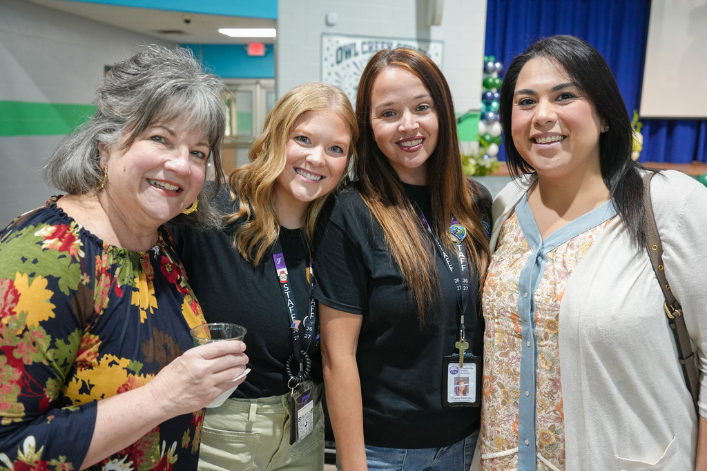 Five women stand together in a brightly lit hallway; one holds a clear cup. A blue curtain is visible in the background.