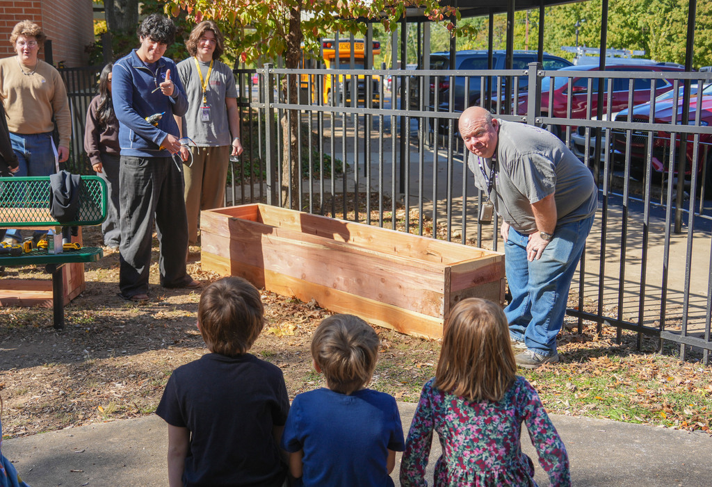 A man shows a large wooden planter to a group of children. The planter is in a paved area.