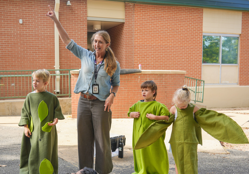 Teacher in light blue shirt and khaki pants speaking with three children in green costumes outside a brick building.
