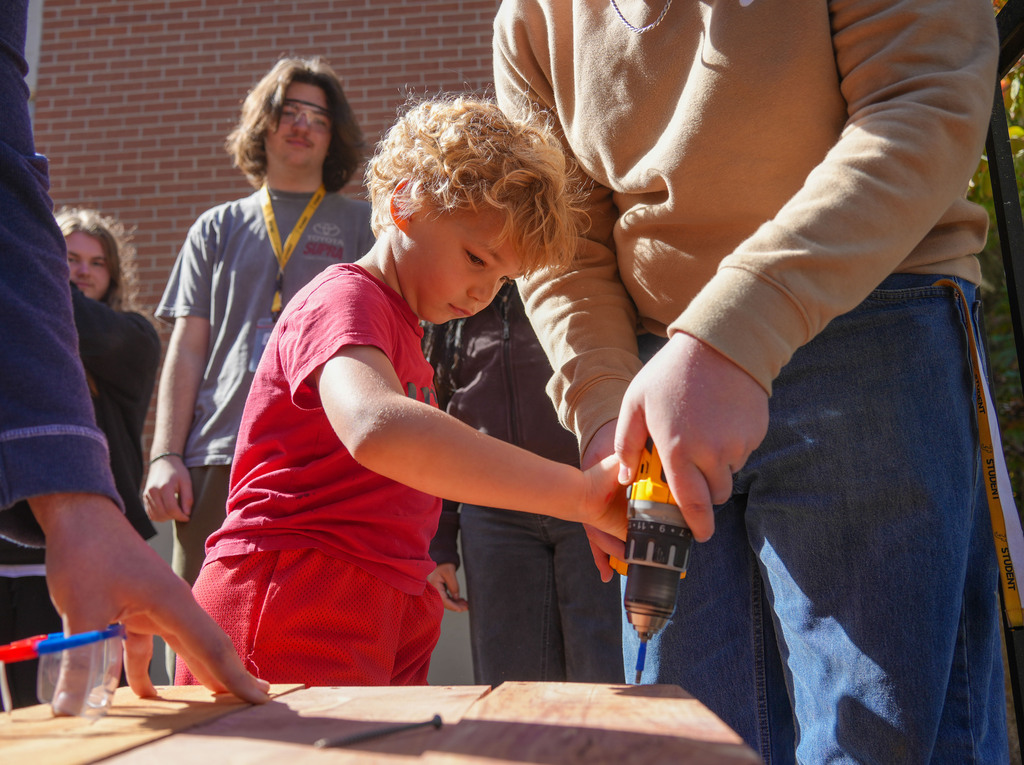 Young child using a drill with help from adult; several people standing behind them watching.