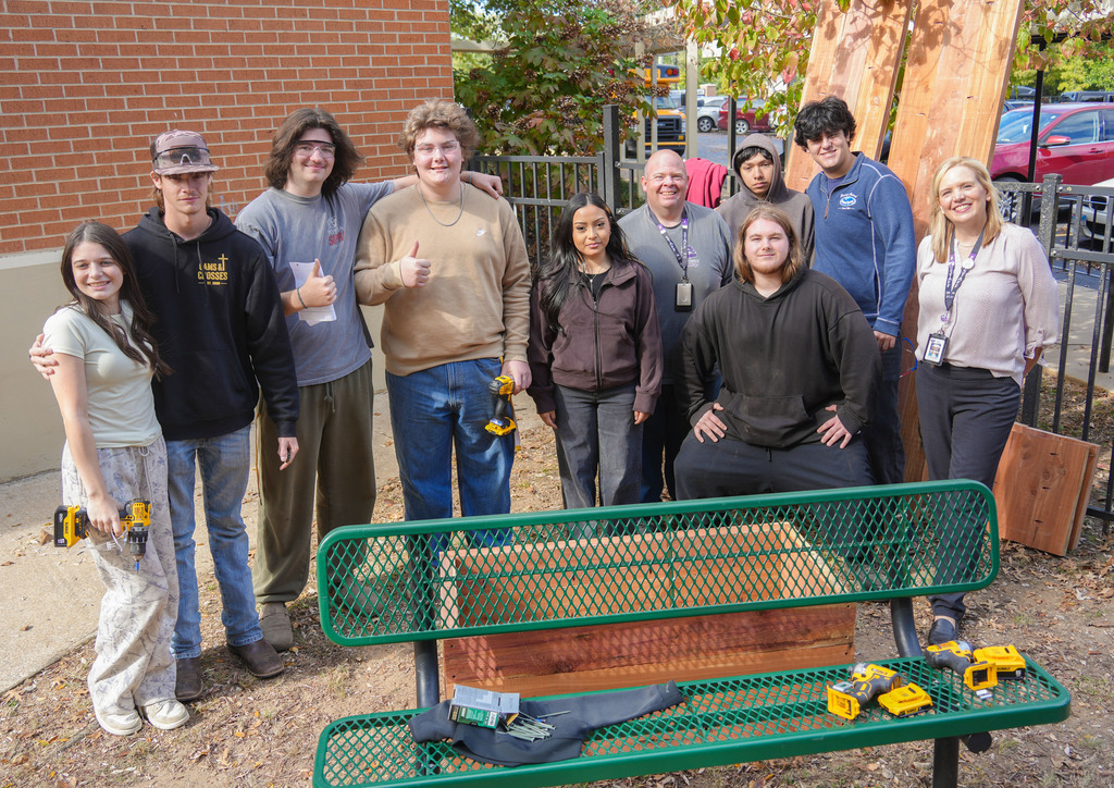 Group of students and adults posing in front of a bench with power tools. Brick building in background.