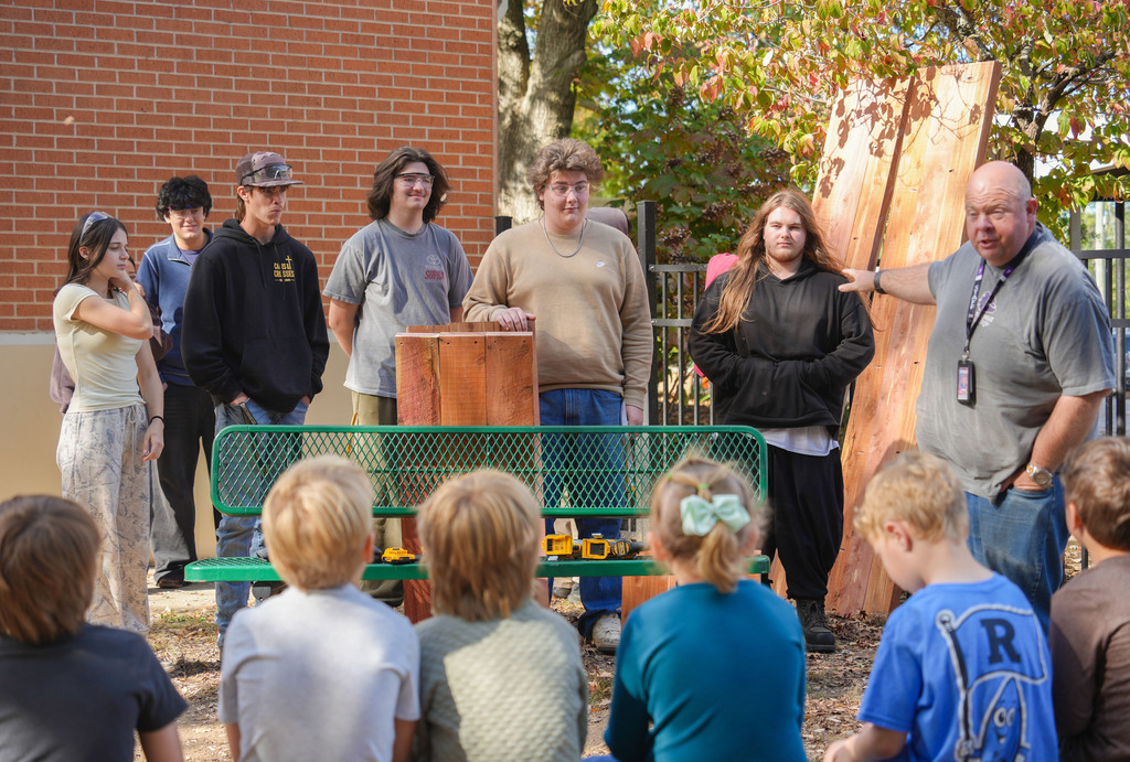 Students and staff standing by a green bench in front of a tree stump.