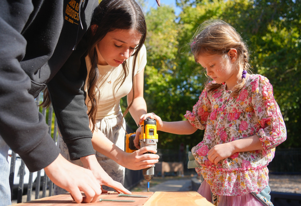 People use tools to work on a wooden surface on a sunny day with trees in the background.