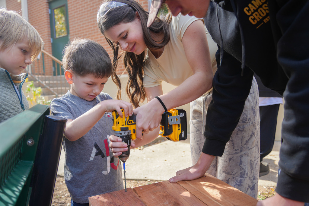 Adult and children together, using a power drill to secure a wooden board. One child holds the drill.