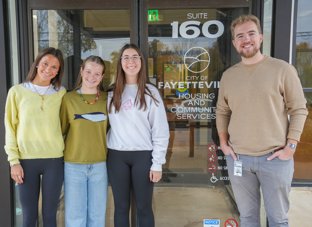 Four young adults stand outside a building. The two in the center wear white shirts and dark pants. The one on the left wears a yellow shirt.