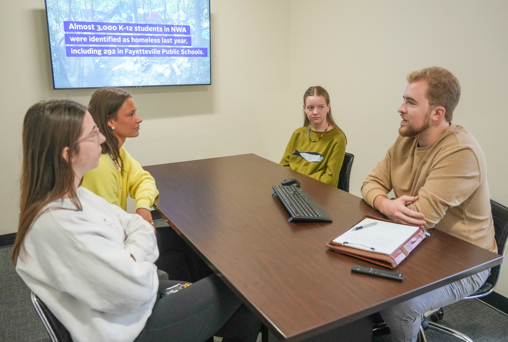 Four people in a conference room sit around a table with a computer, keyboard, and remote.