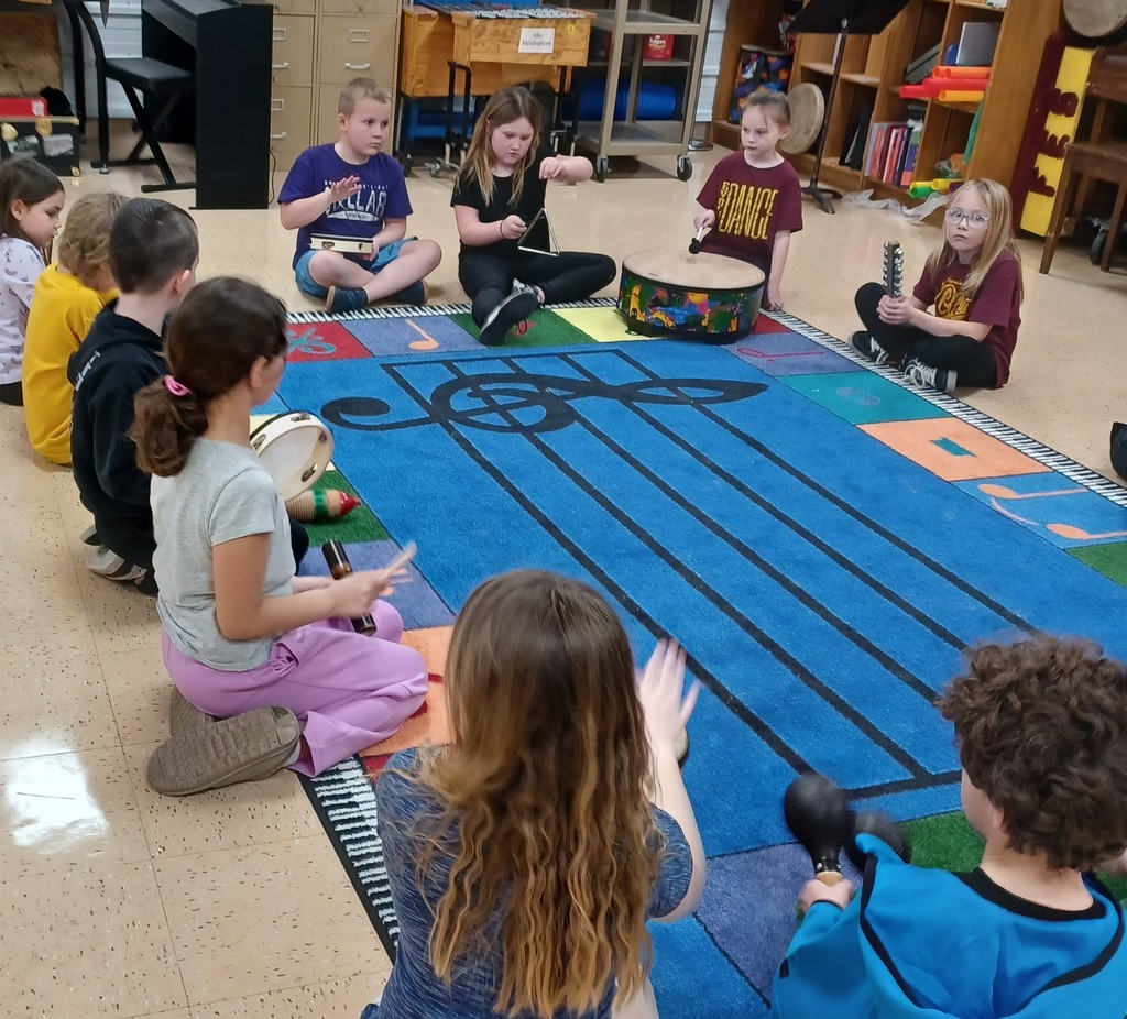 students sit in a circle around a music rug holding different musical instruments