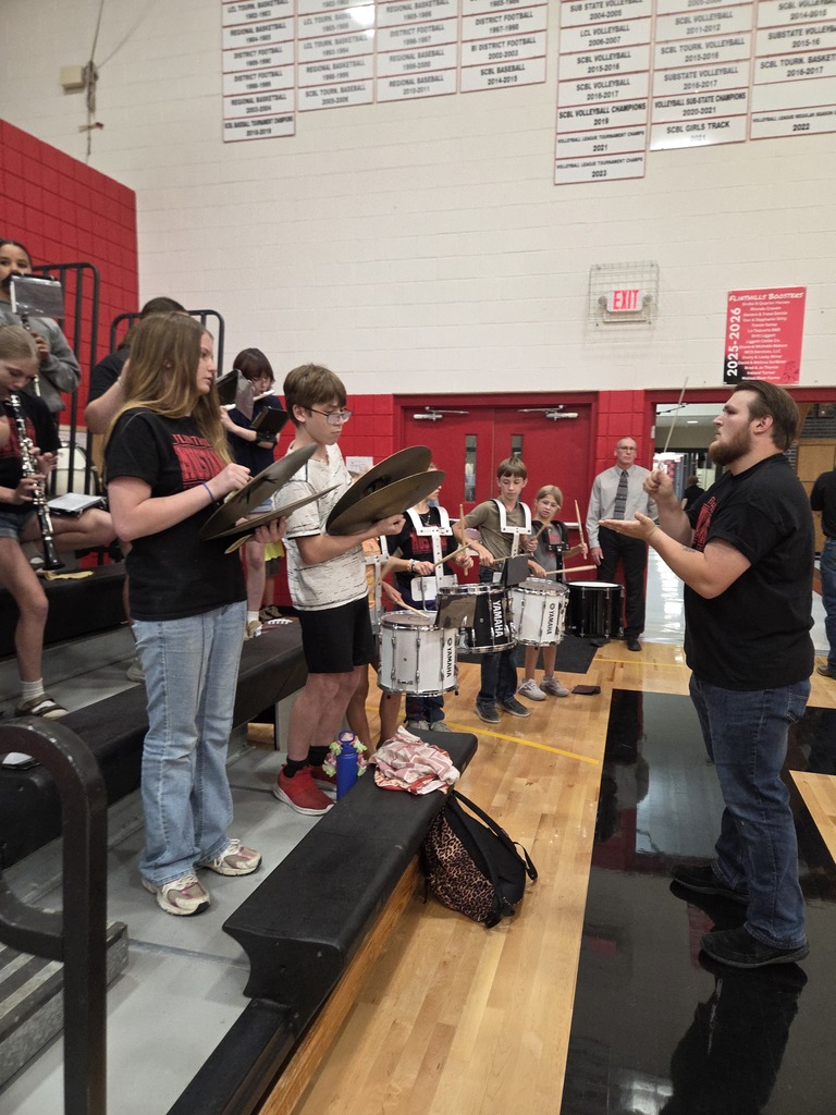 Mr. Green conducts the pep band holding a baton in the school gym at the pep rally. The photo also shows the percussion section.