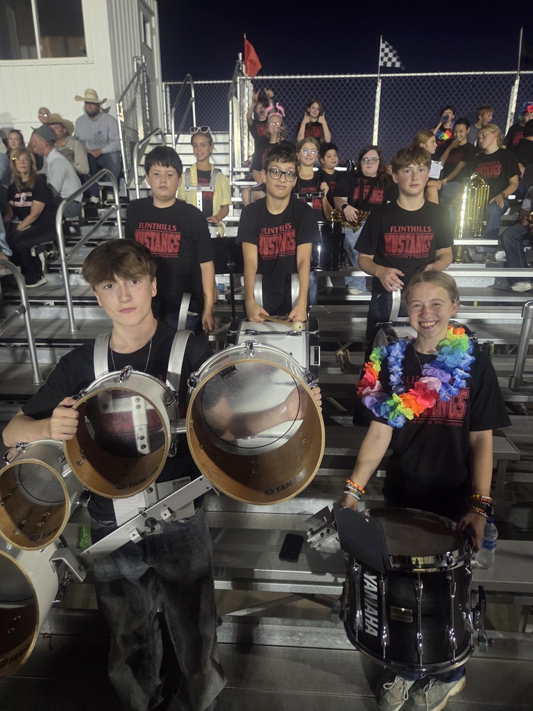 At the center of the photo is five members of the percussion section. In front are a boy and a girl holding Tenor Drums and a Snare respectively. The second row is three boys holding bass drums.