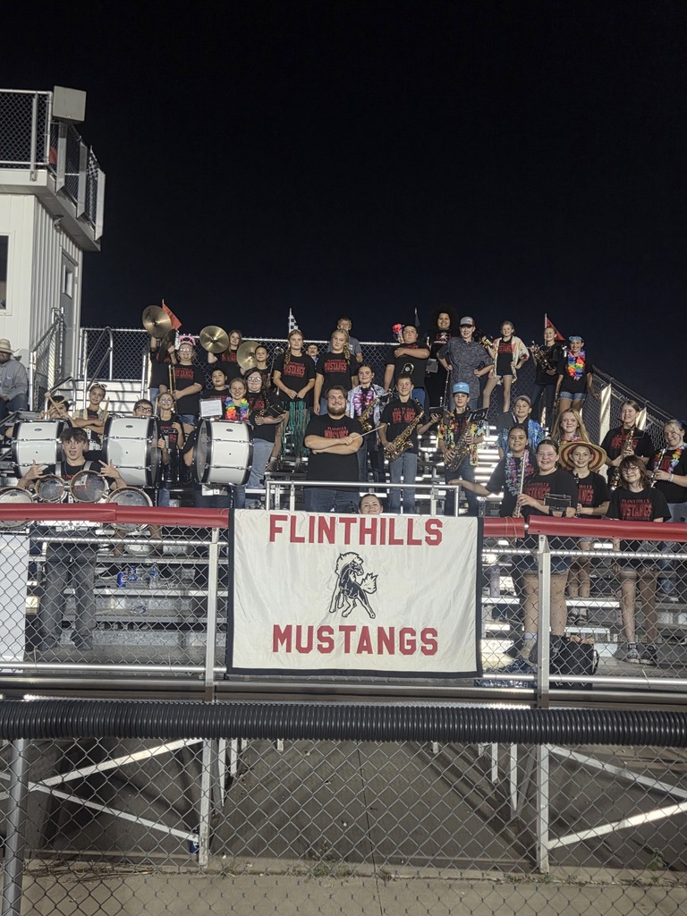 The Flinthills Pep Band stands for a large picture in the football bleachers. A White flag donned with red letters reading "FLINTHILLS MUSTANGS" and the School Mascot are draped over the fencing on the bleachers.