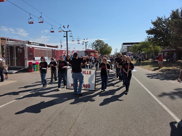 The FHS/FMS bands are marching through the fair grounds as Mr. Davis Green, the Band Director, leads them on. Two student flautists are donning the Flinthills Mustangs banner.