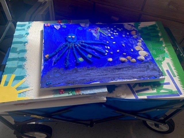 The blue art cart is stacked with canvases. A  blue canvas with a tactile octopus sits atop the cart. A canvas with a yellow sun and  sky of blue squares peeks out from  behind.