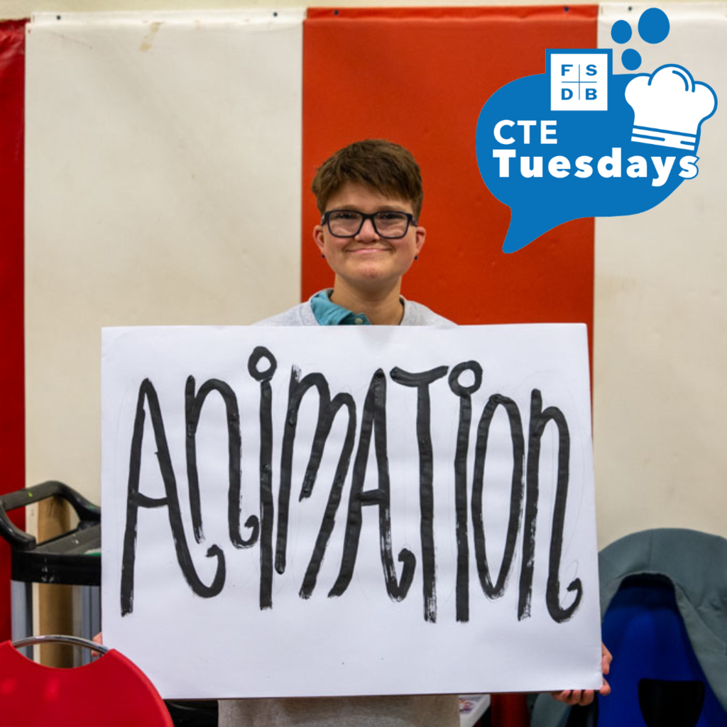 A student is smiling with a big white sign that says "Animation" inside the building where the CTE Showcase was being hosted. 