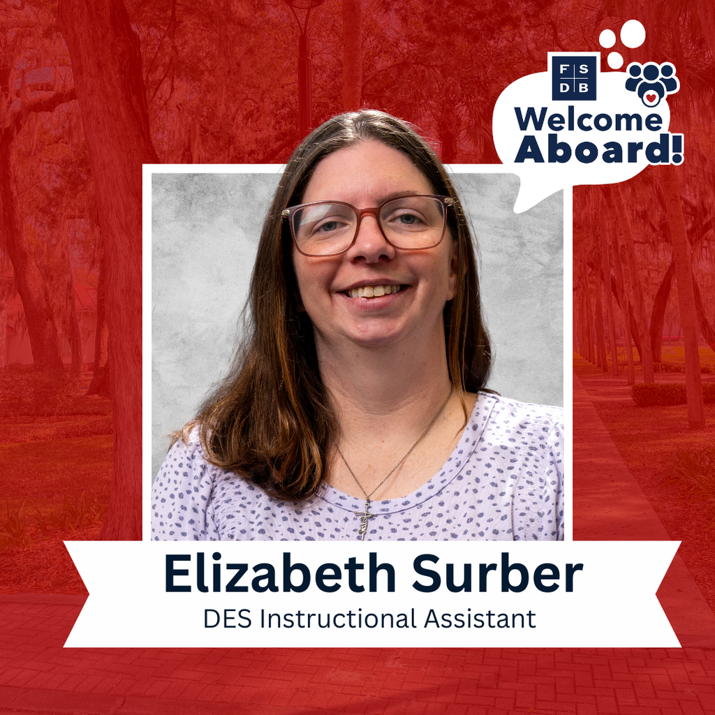 Elizabeth is wearing a light purple with dark purple dots scattered all over shirt is smiling for her photo with a silver backdrop and red background. Below the photo says "Elizabeth Surber, DES Instructional Assistant"