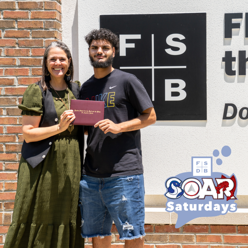 Yeniel is wearing a black shirt with jean shorts holding his diploma with President Snow who is wearing an olive green with black vest next to the FSDB sign.