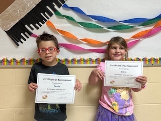 Teddy and Ellie holding their certificates in front of a colorful paintbrush and crepe paper bulletin board