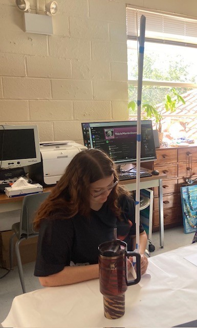 Brooke adds tape to the base of her sculpture. The structure is formed of narrow paper extending straight up from the table in a vertical line, towering above her.