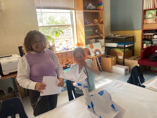 Serenity is holding a piece of paper and smiling for the camera, while Kayla is clapping. Their abstract tower combines an angular base with rolled paper tubes and is topped with a wing-like structure.