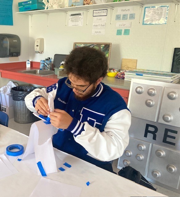 Christian is taping pieces of paper, which are folded lengthwise, onto a triangular base.