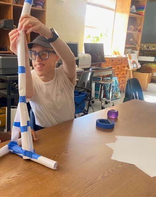 Maddie smiles while adding a paper tube to the top of a tower. The base of the tower has 3 tubes connected to form a tripod.