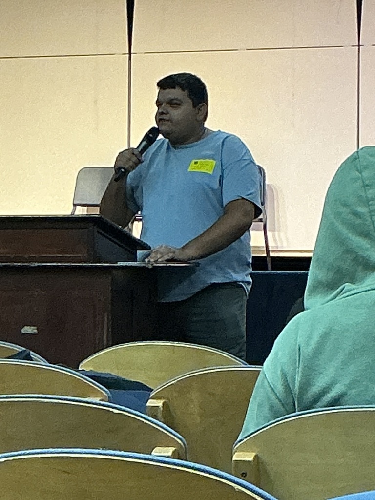 Trent Ferguson stands behind a podium in a blue shirt with a yellow “visitor pass” sticker, holding a microphone. A student in a teal hoodie sits, facing Mr. Ferguson