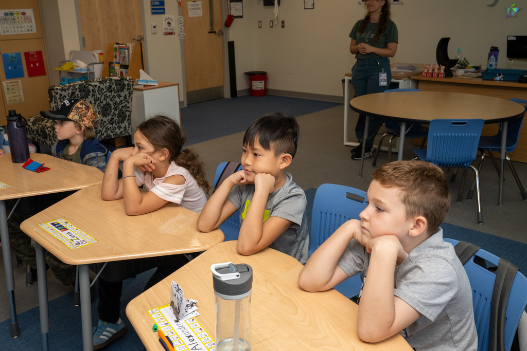 Four students are sitting at their desk watching Brenna sign her book to the class.