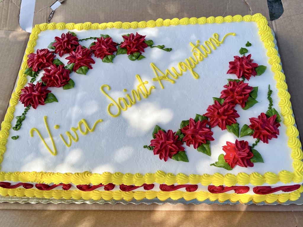 A cake decorated in yellow design, red flowers, and yellow "Viva Saint Augustine".