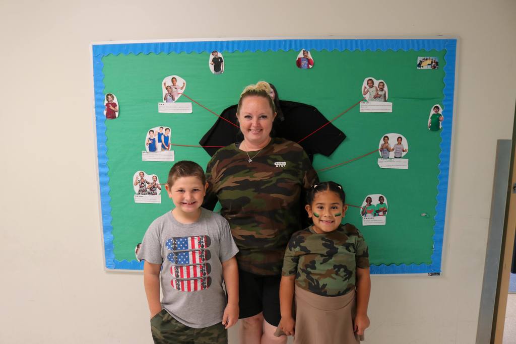 Two students with a staff member dressed in camo smile for a photo in the hallway. 