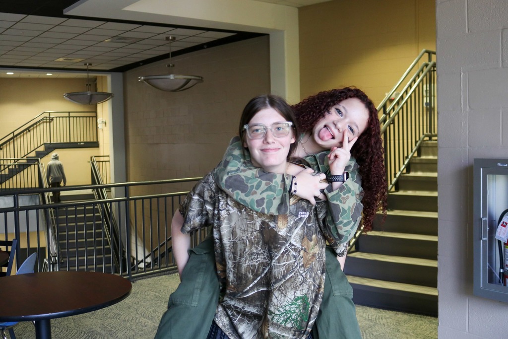 A student giving another student a piggy back ride both wear camo as they smile for a photo in the hallway of Kirk. 