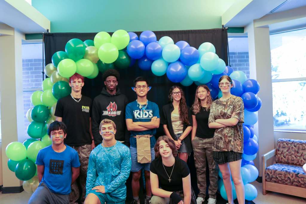 Group of students dressed in camo or blue smile for a photo with green and blue balloon arch. 