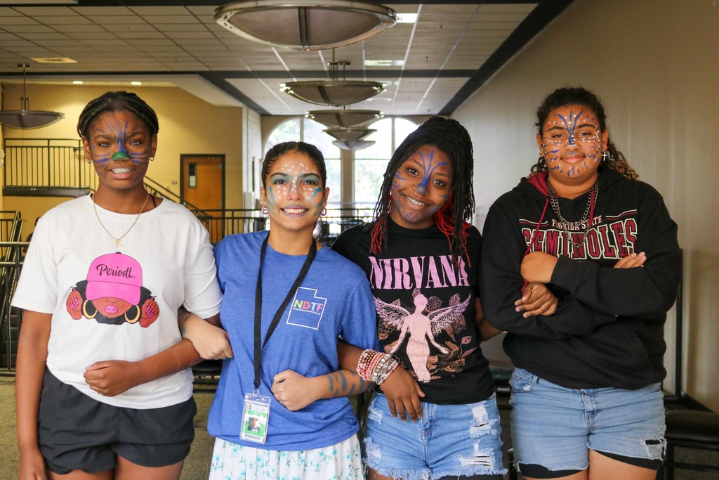 Four students with avatar blue face painting smile for a photo outside the cafeteria. 