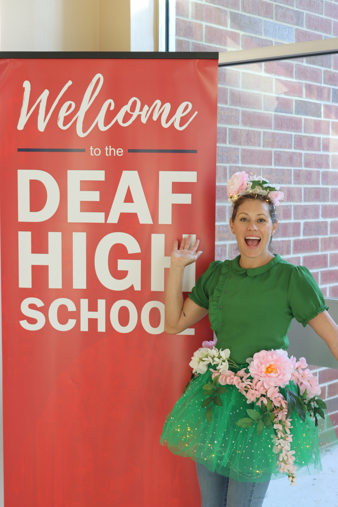 Staff member is wearing a green shirt with green tutu and flowers smiling for her photo next to the "Welcome to Deaf High School" sign. 